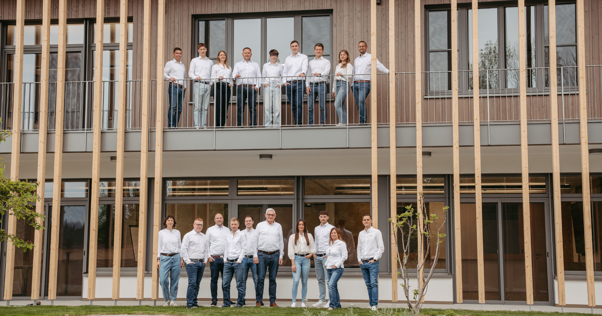 Gruppe von Personen in hellen Hemden und Jeans steht in einer Reihe vor einem großen Stapel Holzstämme.