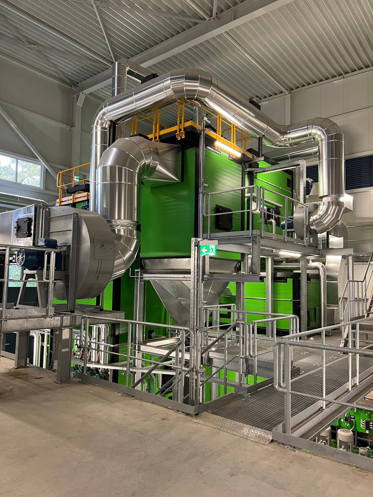 Industrieanlage im Innenraum mit grünem Gehäuse, silbernen Rohrleitungen, Treppen und Geländern; Fluchtweg-Schild sichtbar.