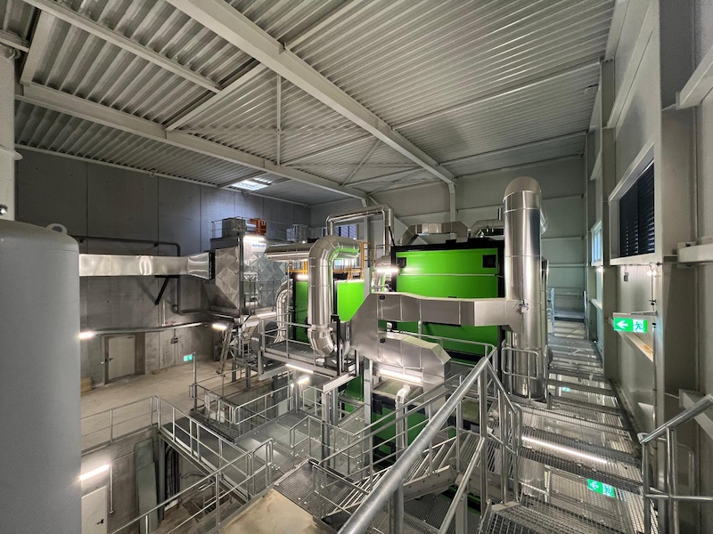 Industrielle Anlage mit grüner Maschine, Metallrohren, Treppengeländern und Laufstegen in einer Fabrikhalle.