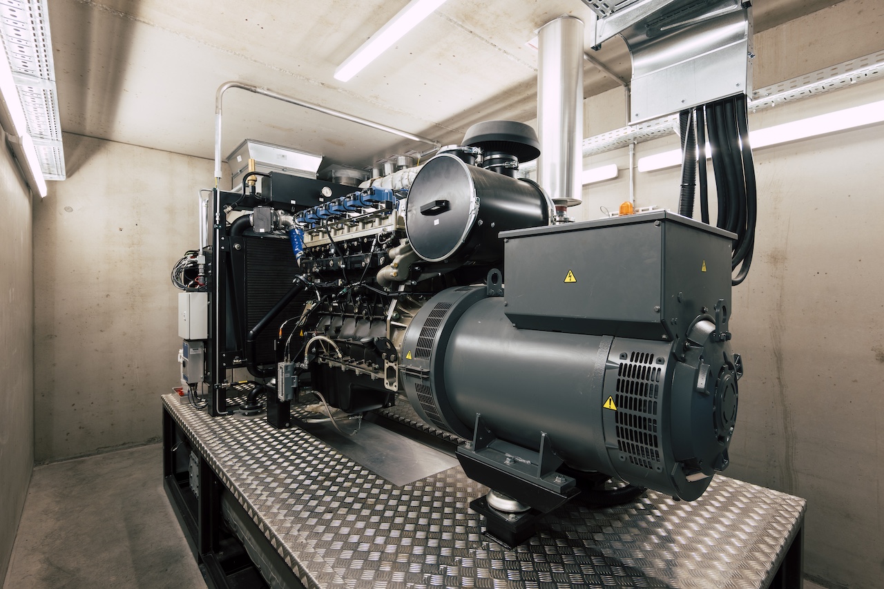 Großer industrieller Generator/Dieselmotor auf Metallplattform in Technikraum, Rohre, Kabel und Lüfter sichtbar.