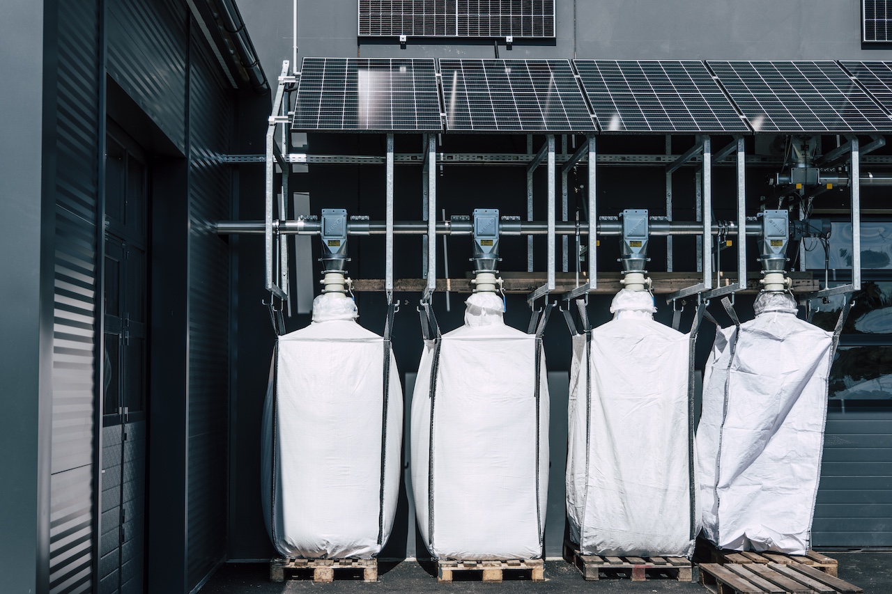 Vier weiße FIBC-Säcke auf Paletten unter einem Rohrleitungs- und Ventilsystem; darüber montierte Solarpaneele.