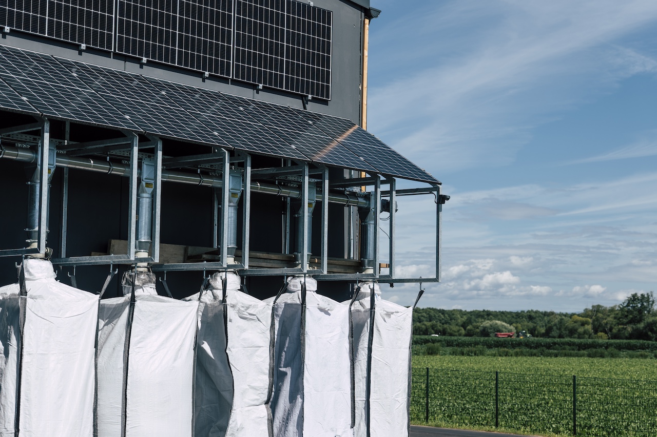 Industriegebäude mit Solarpanelen, Metallkonstruktion und hängenden weißen Abdeckplanen vor grüner Landschaft.