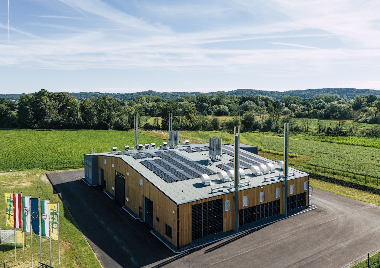 Luftaufnahme eines modernen Holzgebäudes mit Solarpaneelen auf dem Dach, umgeben von Feldern, Bäumen und sanften Hügeln; Flaggen am Rand.