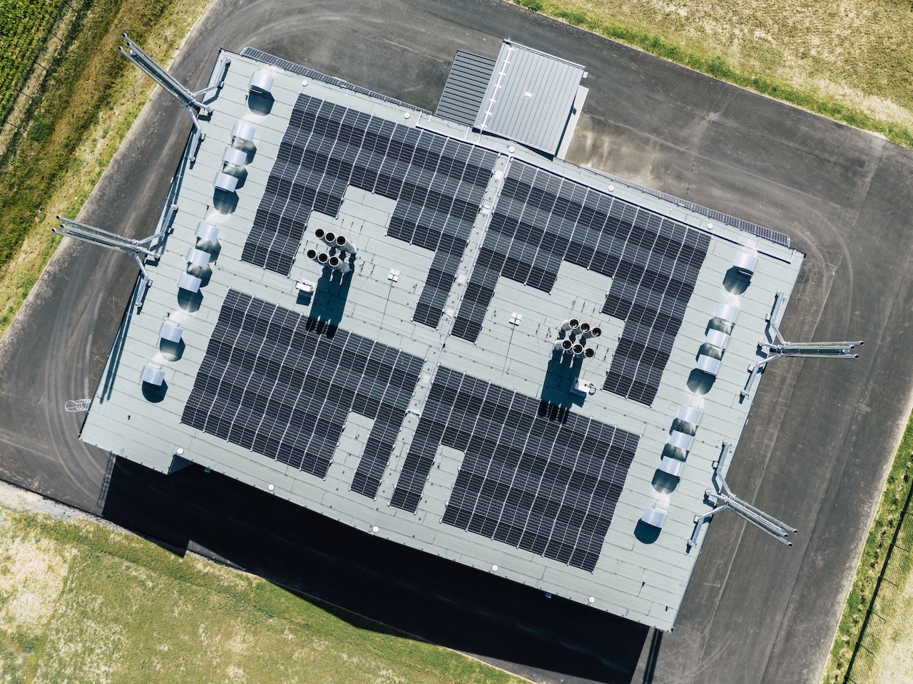 Luftaufnahme eines Flachdachs mit großer Solaranlage aus vielen Modulen und Belüftungselementen; umliegende Wege und Grünflächen.
