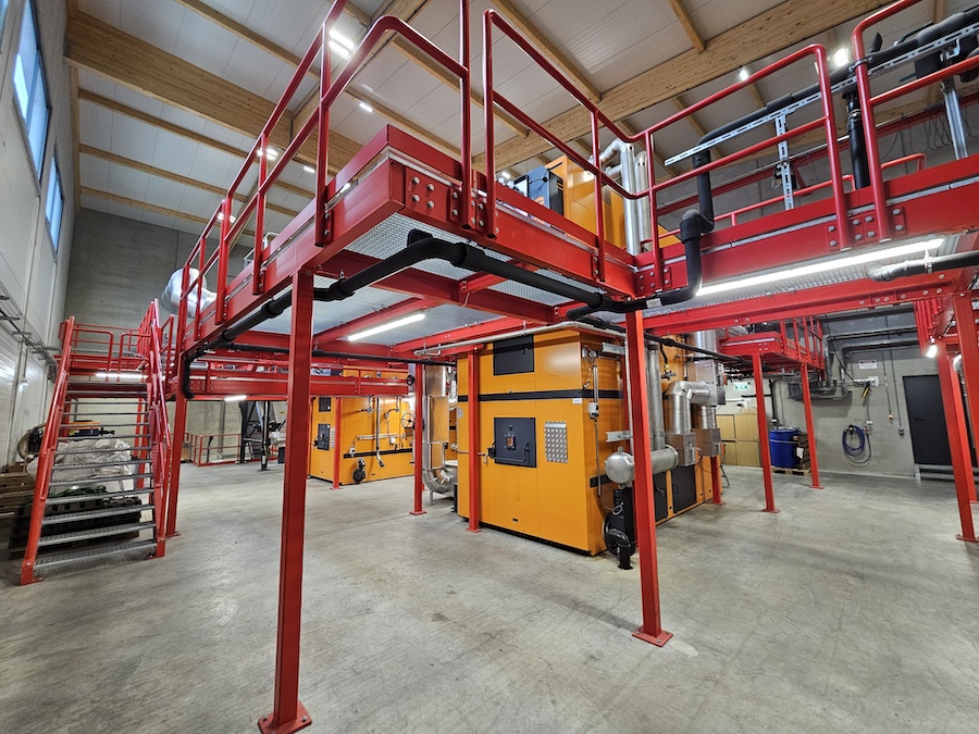 Industrielle Anlage mit rotem Stahlgerüst, Mezzanine, gelb-orange Maschine, Rohrleitungen und Betonboden.