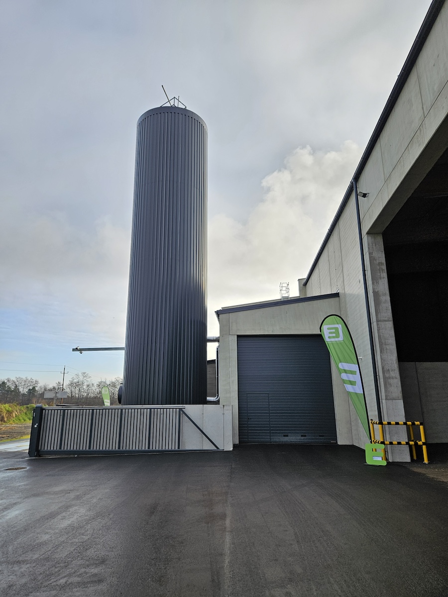 Hoher dunkler Siloturm mit vertikalen Rippen neben einer Halle mit Rolltor; grüne Werbefahne, gelb-schwarz gestreifte Barrieren, nasser Asphalt.