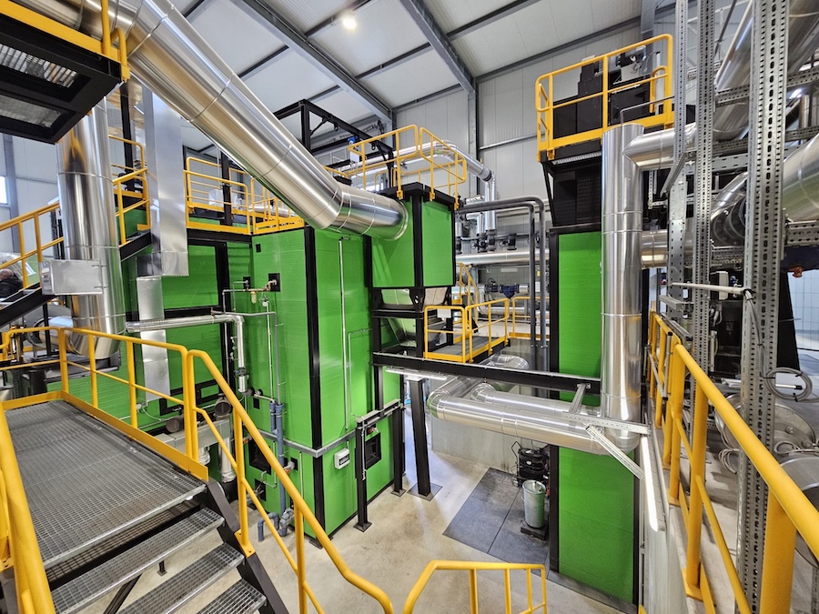 Industrieanlage mit grünen Maschinen, silbernen Rohrleitungen, gelben Geländern und Treppen in einer Fabrikhalle.