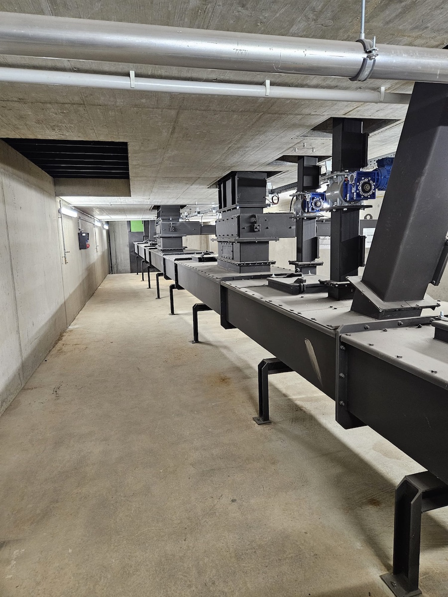 Lange industrielle Förderanlage aus schwarzem Stahl mit Motoren, in einem Beton-Kellerraum; Reihe von Stützen, Laufband und Rohrleitungen.