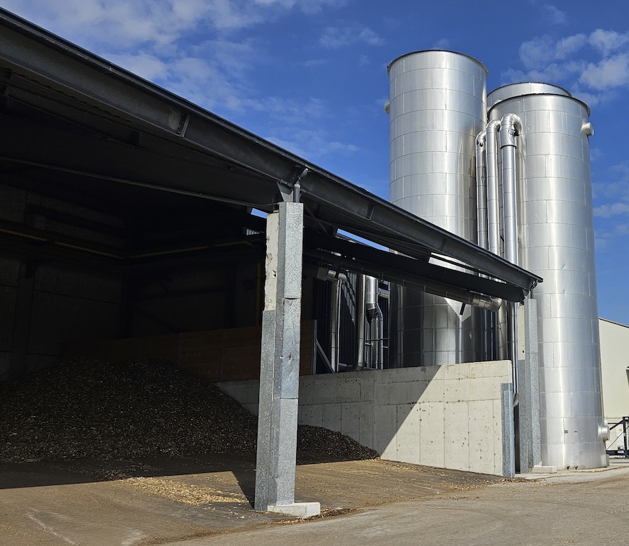 Industrielle Anlage mit zwei großen silbernen Silos rechts, Hackschnitzelhaufen unter Überdachung, Betonmauer, Metallstützen, blauer Himmel.
