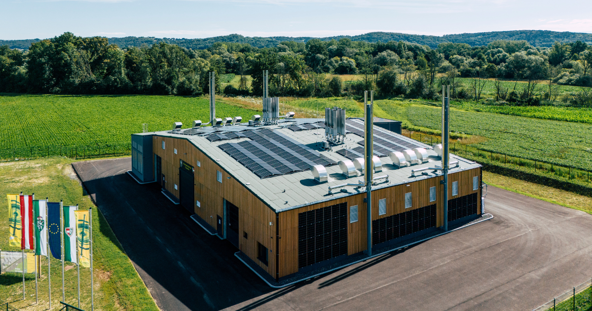 Moderne Holzgebäude-Industrieanlage mit Solarpaneelen auf dem Dach, mehrere Schornsteine, umgeben von Feldern und Wald; Flaggen am Eingang.