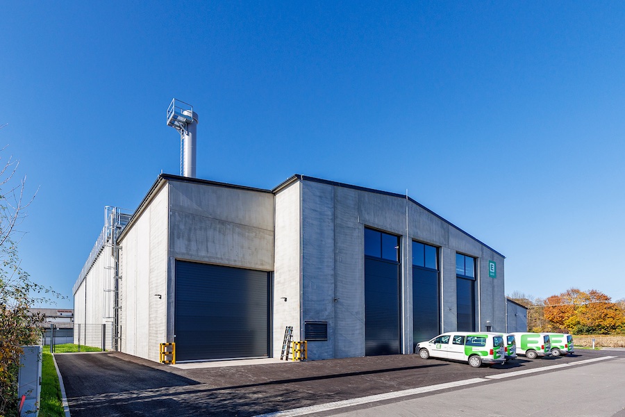Beton-Lagerhalle mit drei großen Rolltoren; davor parkende weiße Firmenwagen, blauer Himmel und herbstliche Bäume am Rand.