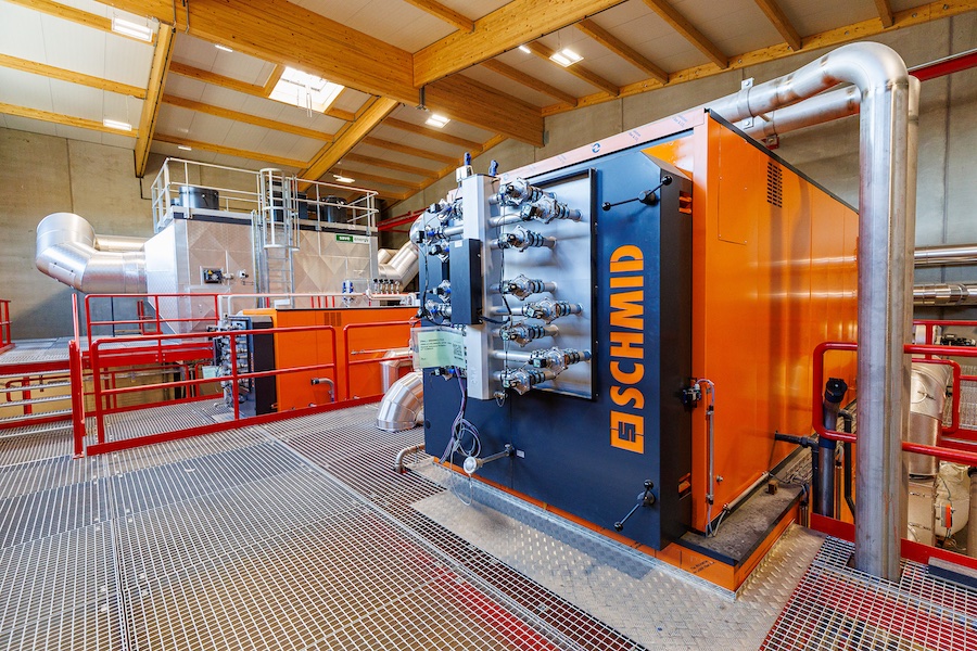 Industrielle Anlage mit orange-blauem Maschinenblock, Rohrleitungen und Metallgitterboden.