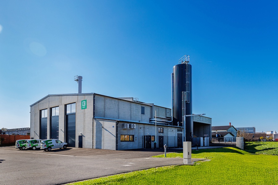 Industrieanlage mit großer Halle, rechts stehender Siloturm, Parkplätze mit Firmenfahrzeugen, grünem Rasen und blauem Himmel
