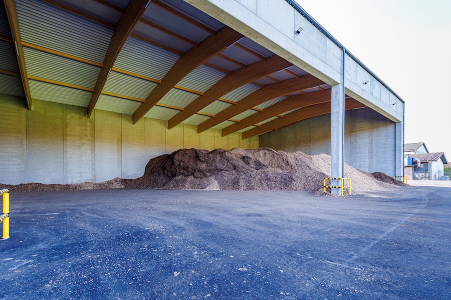 Große überdachte Lagerhalle mit Haufen Holzspäne auf Asphalt; Holzbalkendecke, gelbe Sicherheitspfosten, offener Seitenbereich.