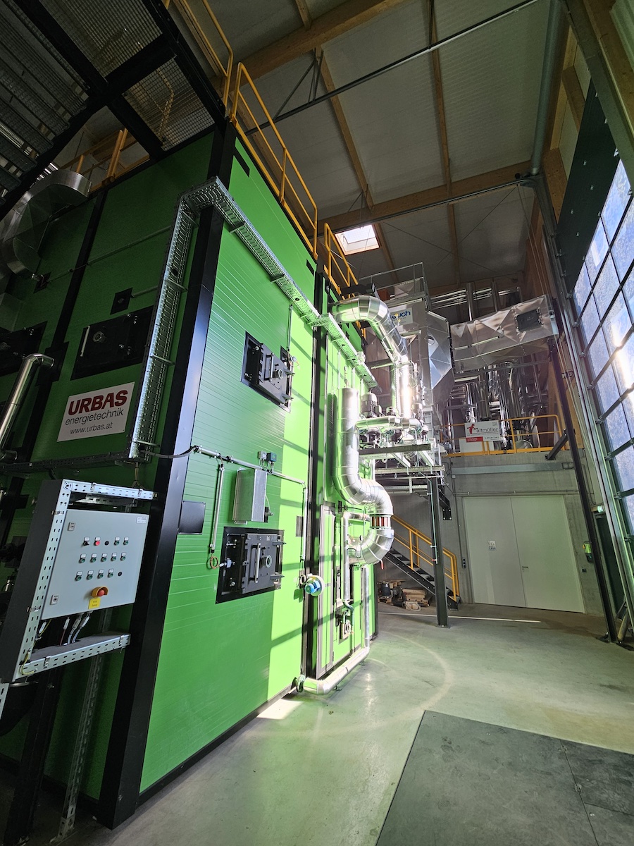 Große grüne Industrieanlage mit silbernen Rohrleitungen, Geländern und Treppen in einer Fabrikhalle.