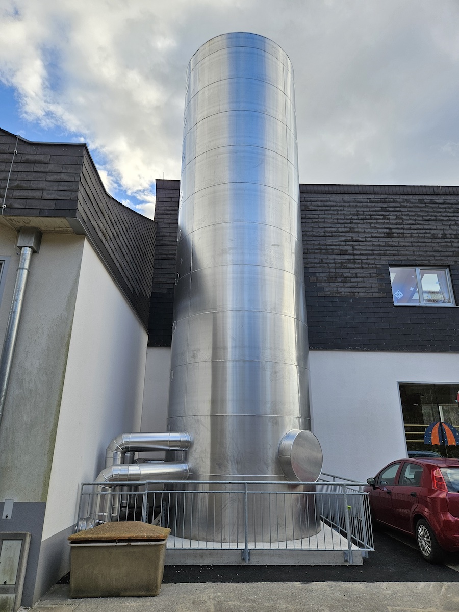 Großer silberner Pufferspeicher im Außenbereich eines Gebäudes, mit Leitungen, Geländer, Müllcontainer und rotem Auto.