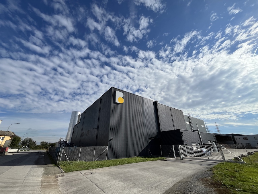 Dunkles Industriegebäude aus Metall mit gelbem Logo, Zaun und Zufahrt, unter blauem Wolkenhimmel.