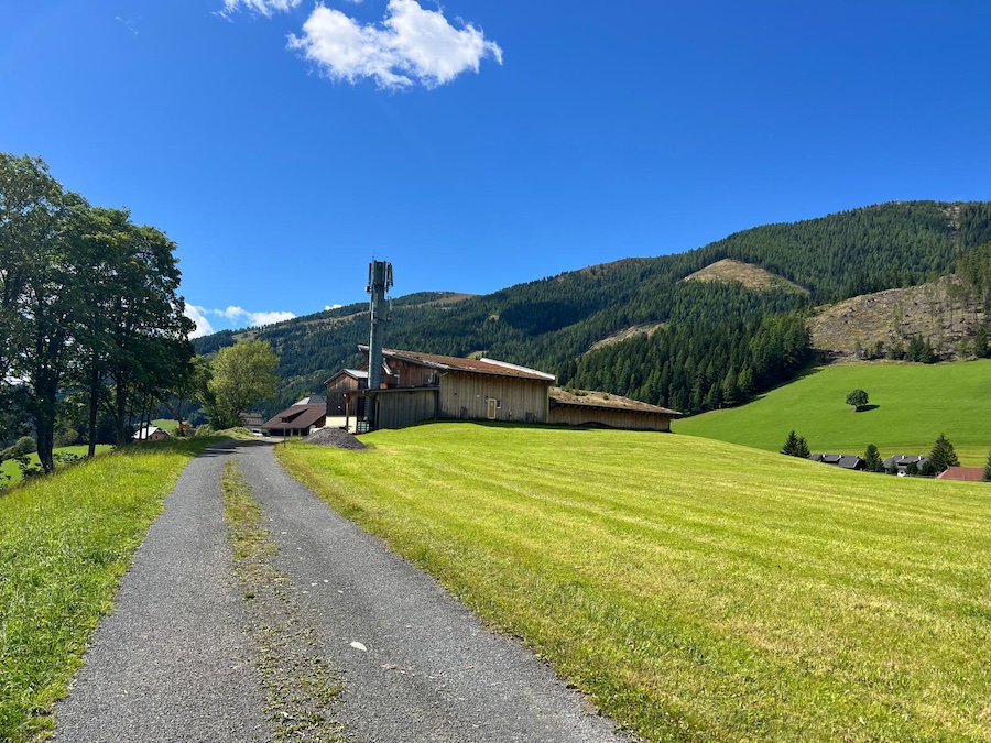 Ländliche Landschaft: schmale Asphaltstraße durch grüne Wiesen führt zu Holzbauten und einem Funkmast; im Hintergrund Waldberge unter blauem Himmel.