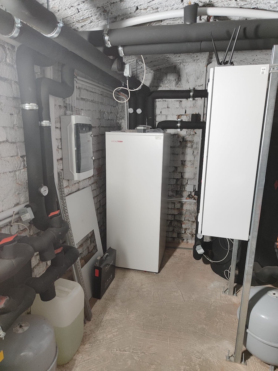 Keller-Heizungsraum mit weißen Wärmeerzeugern, schwarzen Rohren, Pumpen und Messgeräten vor Bruchsteinwand.