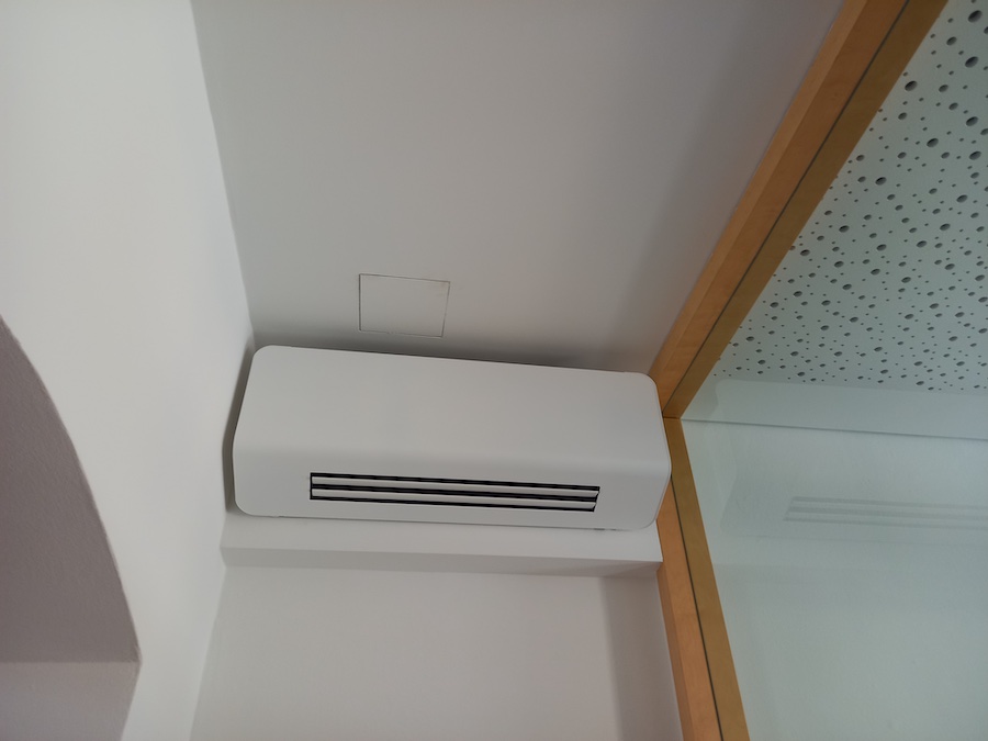 Weißes Wandgerät mit horizontalen Lüftungsschlitzen, vermutlich Klimaanlage, an der Wand nahe Decke installiert; rechts Glaswand mit Holzrahmen.