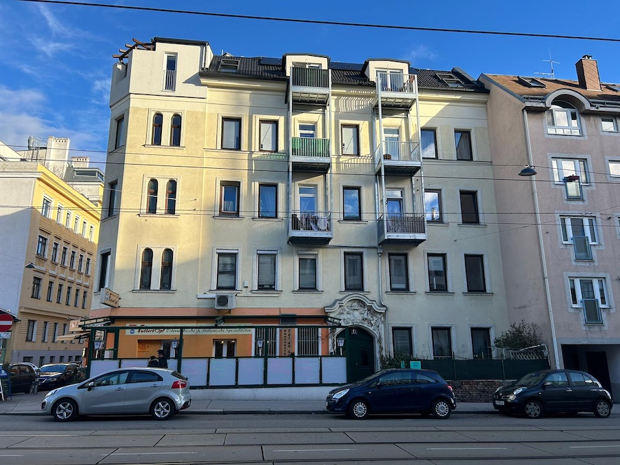 Mehrstöckiges Wohngebäude mit Balkonen, Erdgeschossgeschäft, davor parkende Autos und Straßenbahngleise vor blauem Himmel.