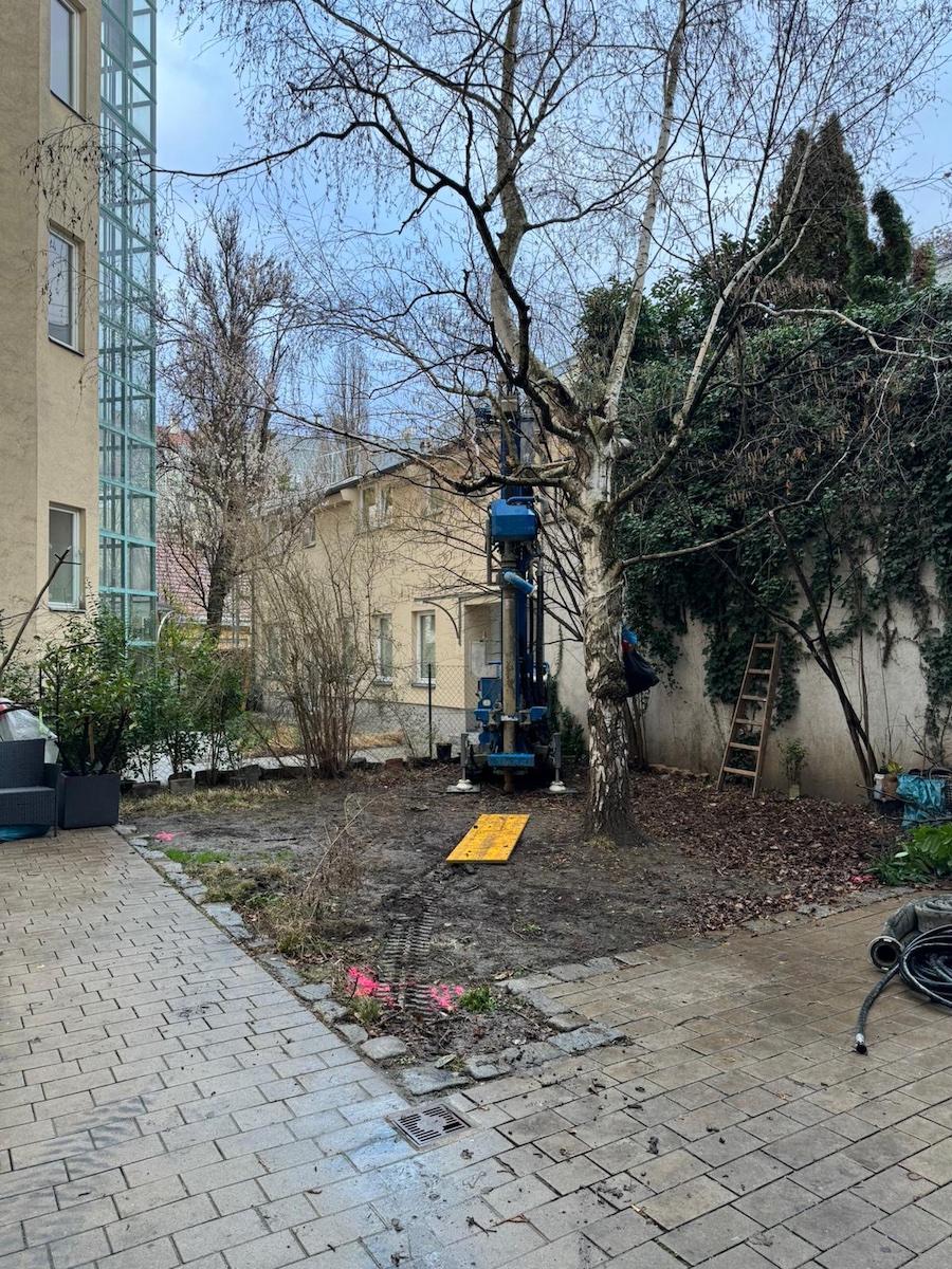 Blaues Bohrgerät neben Baum in Innenhof; Leiter, Kabel und gepflasterte Hoffläche sichtbar.
