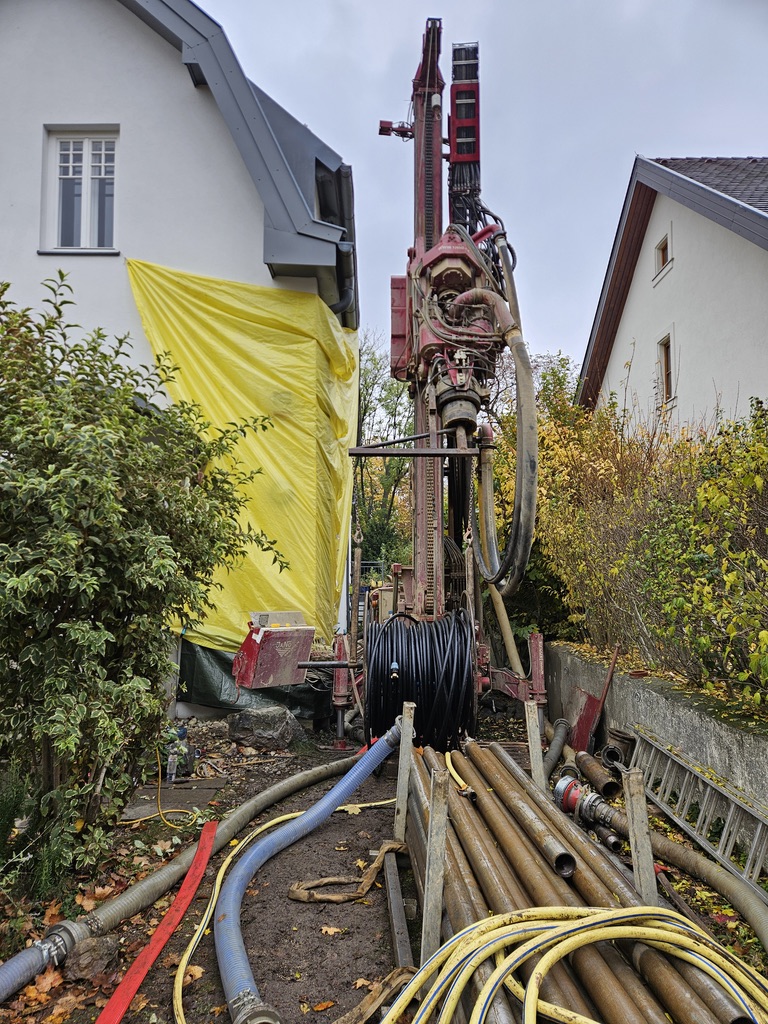 Bohrgerät auf einer Baustelle zwischen zwei Häusern, umgeben von Schläuchen, Rohren und einer gelben Abdeckung.