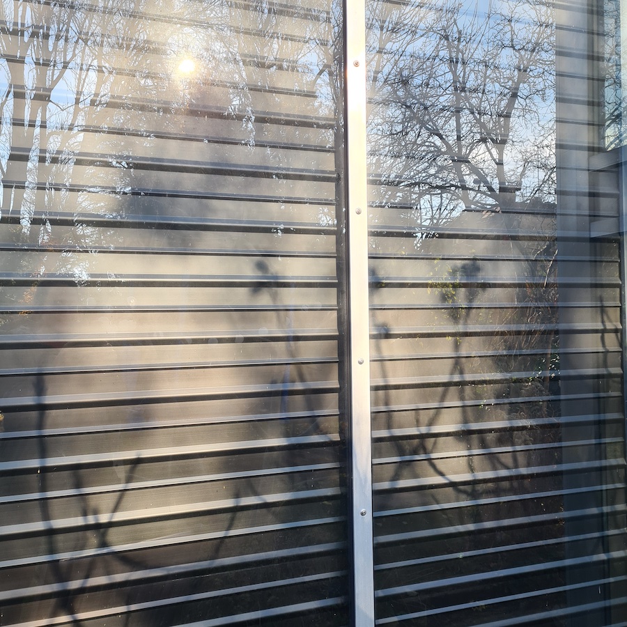 Solarthermie Hausfront mit horizontalen Lamellen und Metallrahmen; Reflektionen von Bäumen und Sonnenlicht im Glas.
