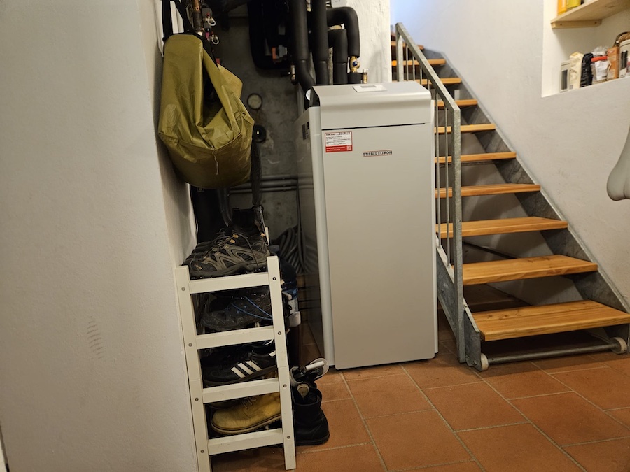 Kellerraum mit Treppe rechts, Holzstufen und Metallgeländer; Schuhregal mit Schuhen, olivgrüner Beutel an der Wand, weißes Hochgerät, Fliesenboden.