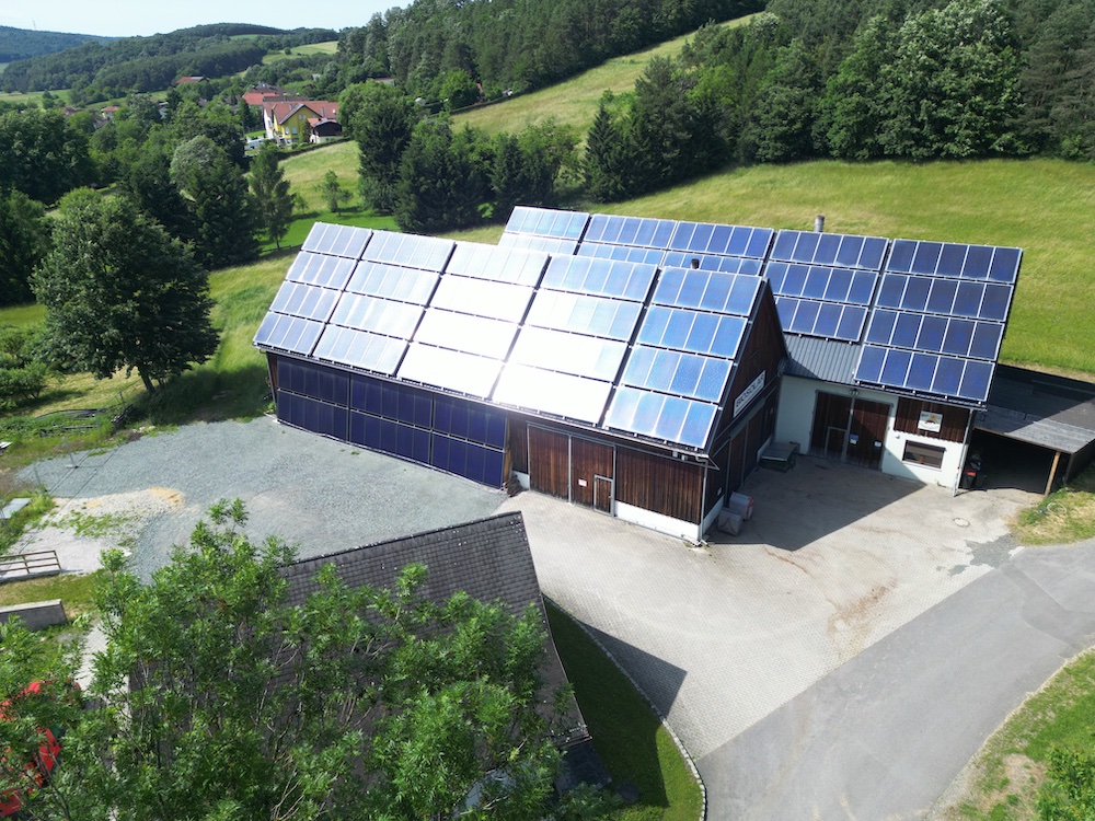 Ländliches Gebäude mit Solarpaneelen auf dem Dach, umgeben von Bäumen und Hügeln.
