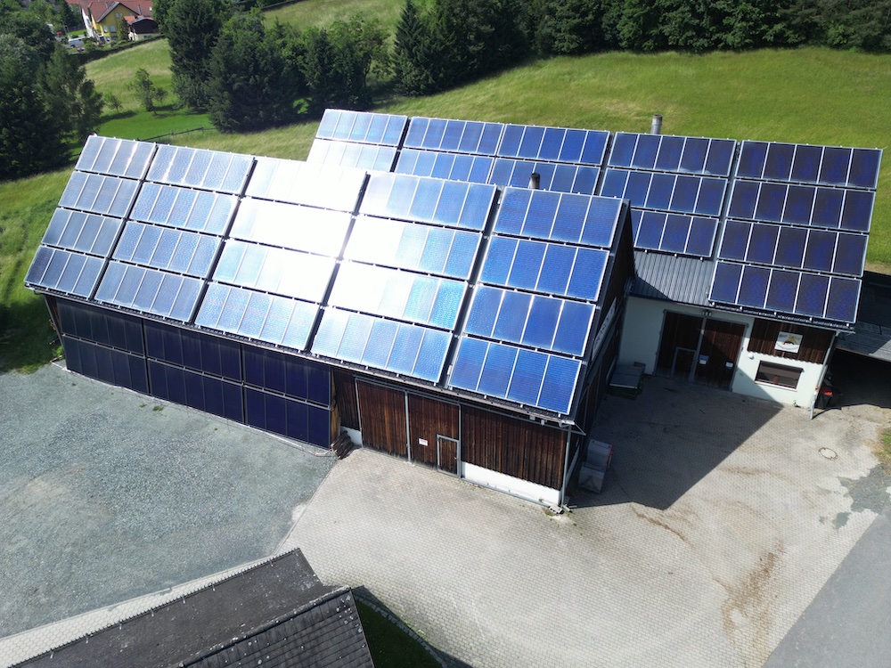 Gebäude mit vielen Solarmodulen auf dem Dach; grüne Wiesen und Bäume im Hintergrund.