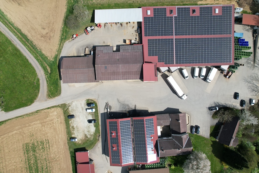 Luftaufnahme eines Firmengeländes mit Solaranlagen auf Dächern, mehreren LKWs, Gebäuden und umliegenden Feldern.