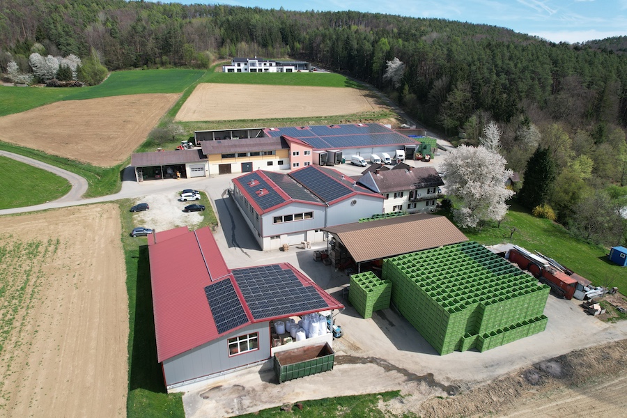 Luftbild eines landwirtschaftlichen Betriebs mit Solardächern, Lagergebäuden, großen grünen Stapelkisten und umliegenden Feldern.