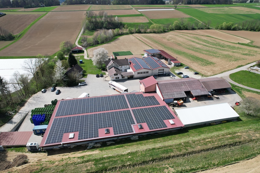 Luftaufnahme eines Hofgeländes mit Solardächern, Gebäuden, Fahrzeugen, Parkplätzen und umliegenden Feldern.