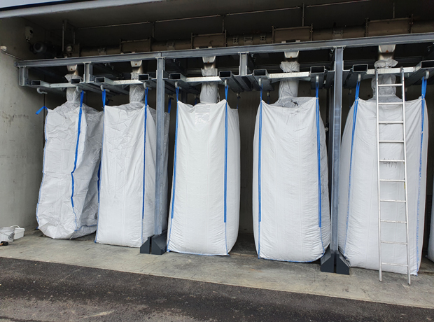 Mehrere große weiße FIBC-Säcke mit blauen Bändern hängen unter einem Metallträger; rechts eine Leiter, Boden aus Beton.