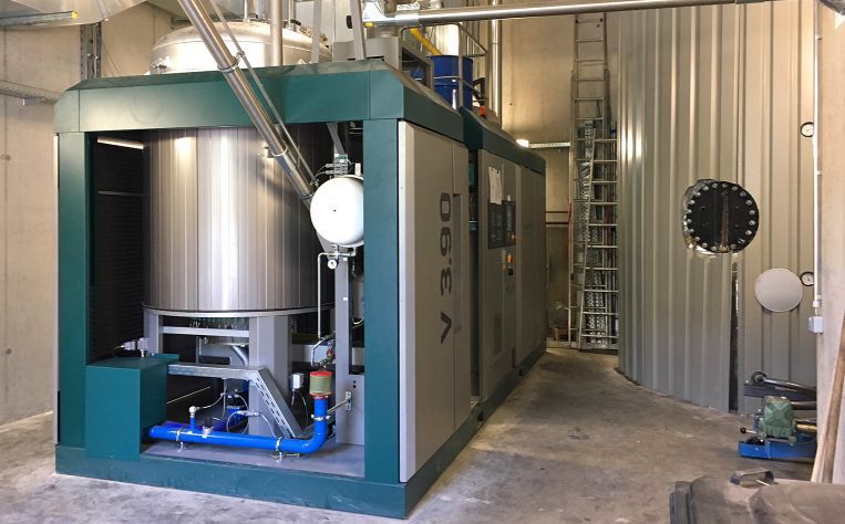 Große industrielle Anlage in einer Halle mit grün-silbernem Gehäuse, Rohrleitungen, Bedienelementen und Leiter im Hintergrund.