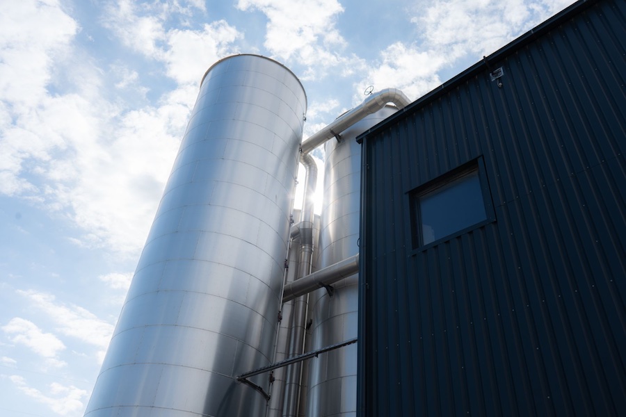 Großes silberner Pufferspeicher mit Rohrleitungen neben einem dunklen Stahlgebäude vor blauem Himmel mit Wolken.