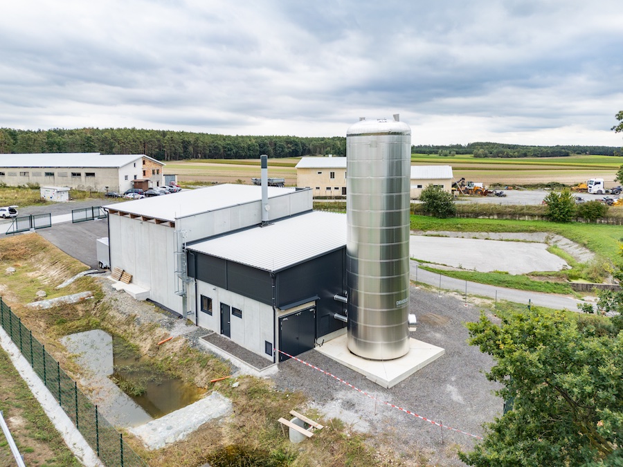 Außenansicht einer Industrieanlage mit großem silbernen Metallsilo, Bürogebäuden und umliegenden Feldern.