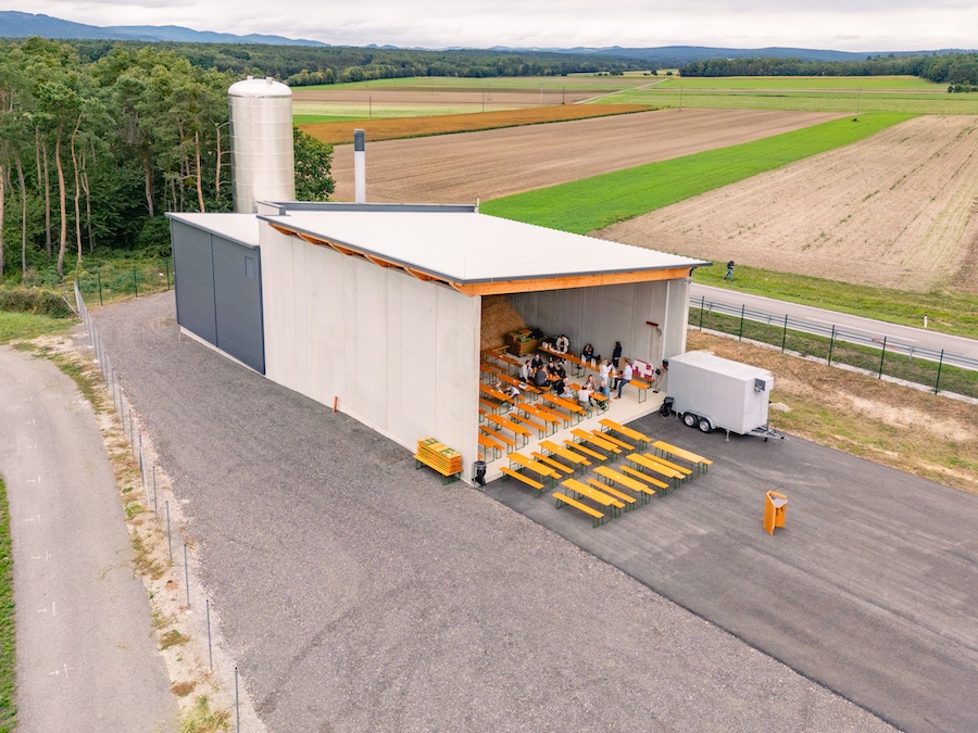 Offene Scheune mit gelben Tischen und Bänken; Menschen sitzen an Tischen, daneben Anhänger, Felder und Straße im Hintergrund.