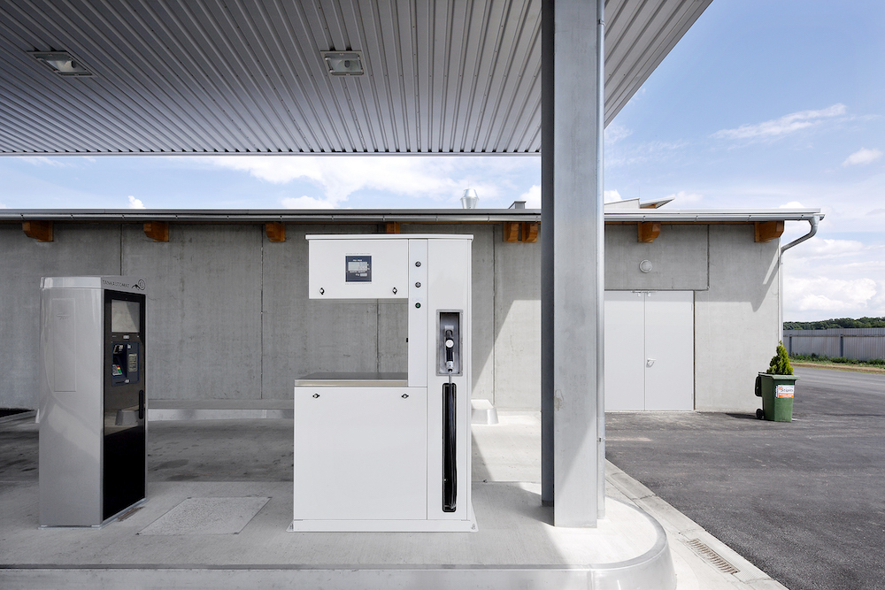 Unter Überdachung befindliche Tankstelle mit Zapfsäule, digitalem Display, zentralem Automatengehäuse und Betonwand.