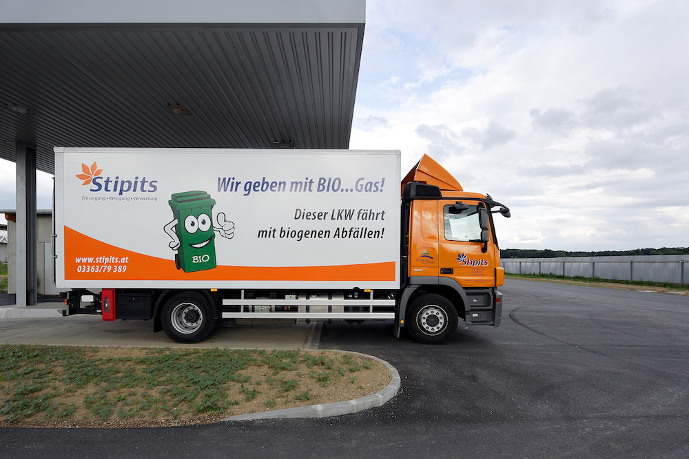 LKW mit orangefarbenem Fahrerhaus und weißem Auflieger der Firma Stipits; BIO-Gas-Werbung und biogene Abfälle, Cartoon-Bin-Illustration.