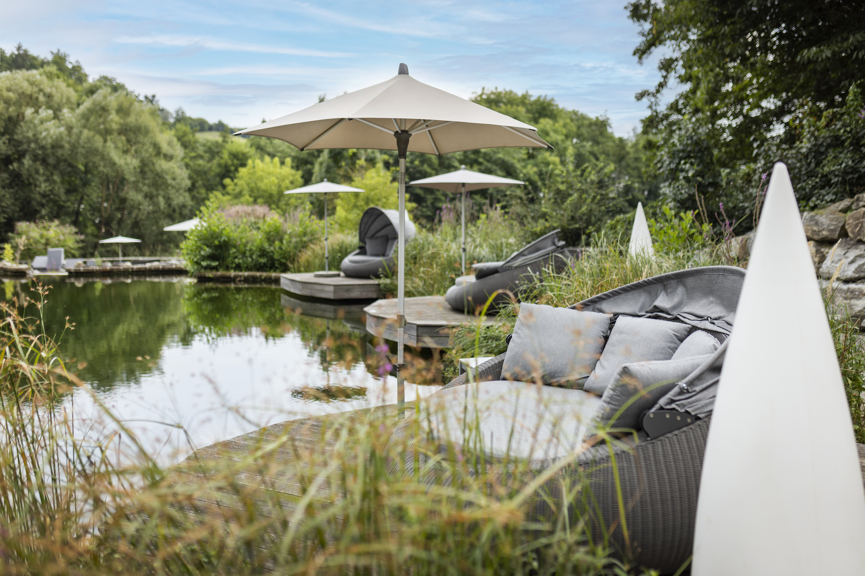 Loungebereich am Teich mit Sonnenschirmen, Rattanmöbeln, Kissen und grüner Gartenlandschaft.