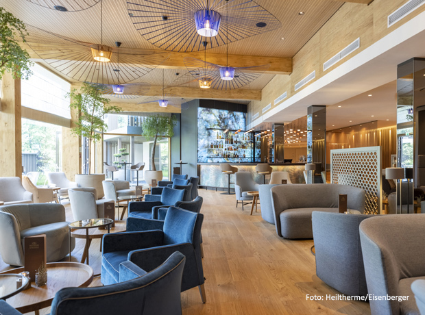 Heller Lounge-Bereich mit Holzboden und Balkendecke, blaue Sessel, graue Sofas, Bar im Hintergrund und Grünpflanzen.