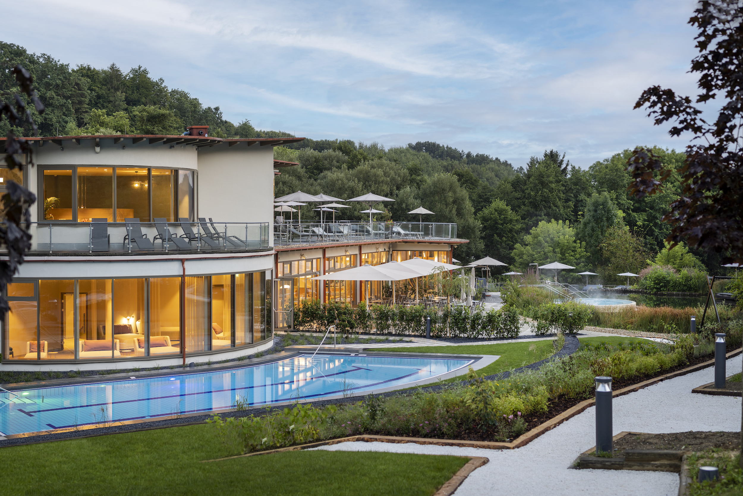Modernes Resortgebäude mit großen Glasfronten, warm beleuchtetem Innenbereich, Terrasse mit Liegen und Sonnenschirmen, Außenpool und Garten.