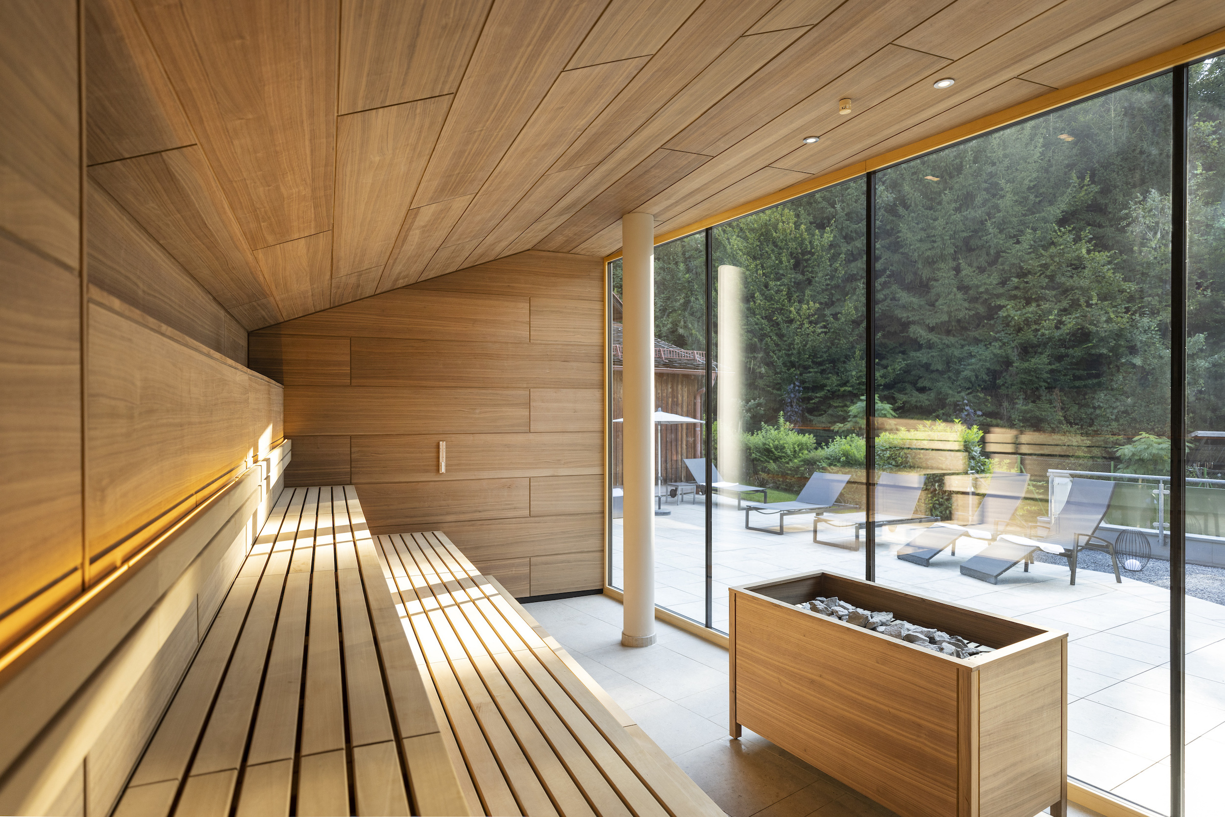 Moderne Holzsauna mit langen Bänken und Glasfront, Ofen mit Steinen; Außenbereich mit Liegen und grünem Garten sichtbar