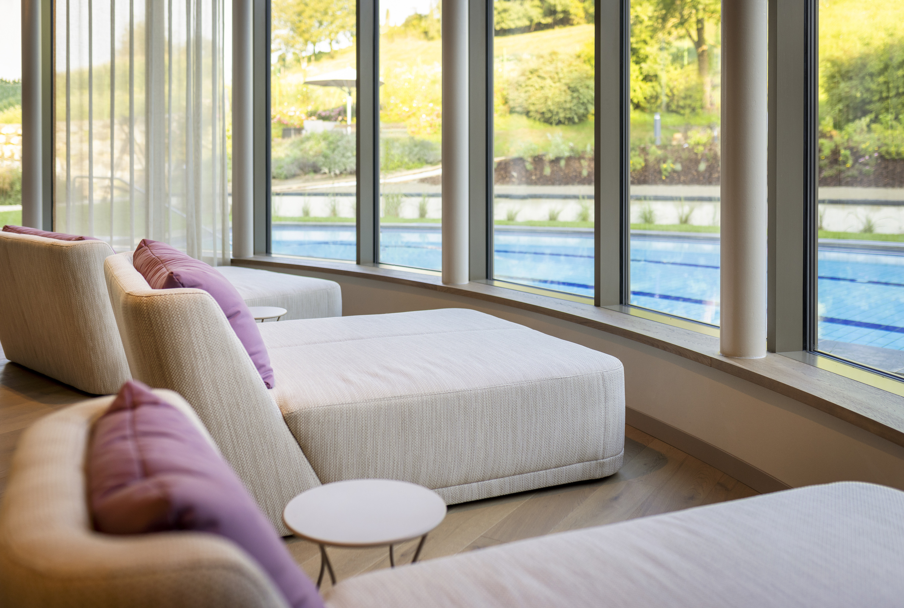 Heller Loungebereich mit beigen Liegen und lila Kissen, Fensterfront mit Blick auf Pool und Gartenlandschaft.