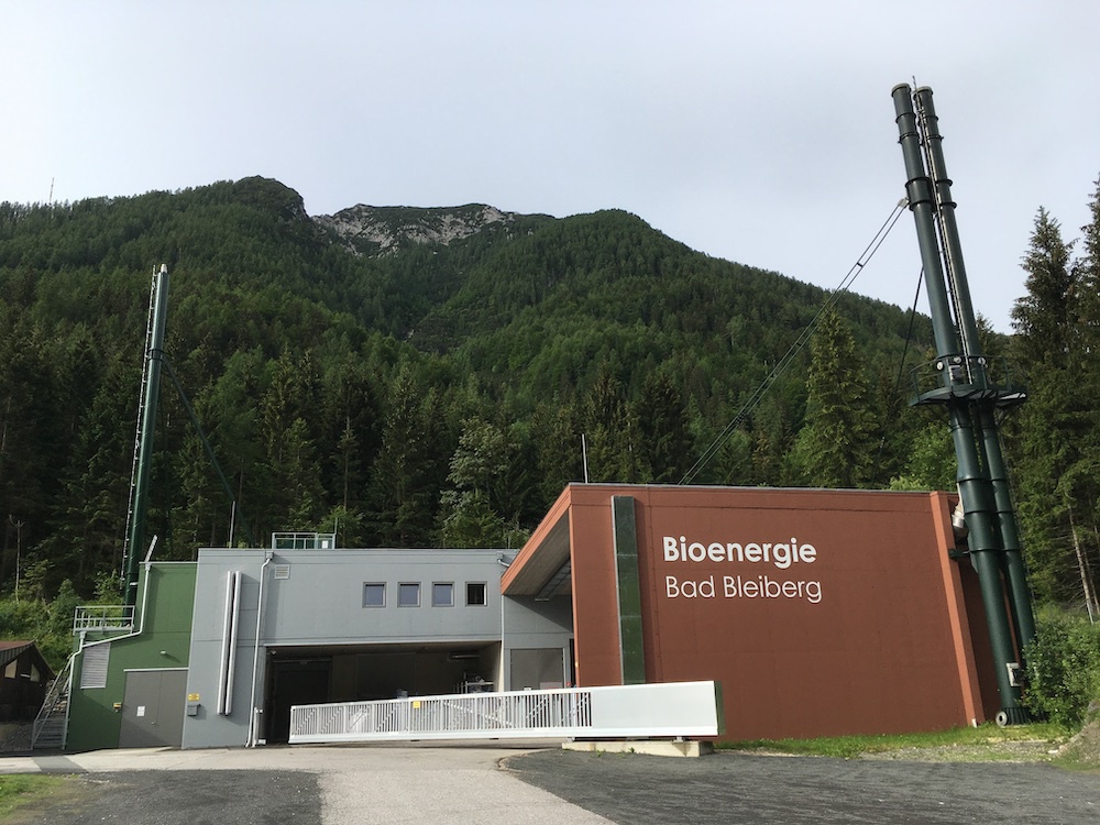 Bioenergie Bad Bleiberg: Moderne Anlage am Waldrand mit brauner Fassade und hohen Turmrohren vor Wald- und Bergkulisse.