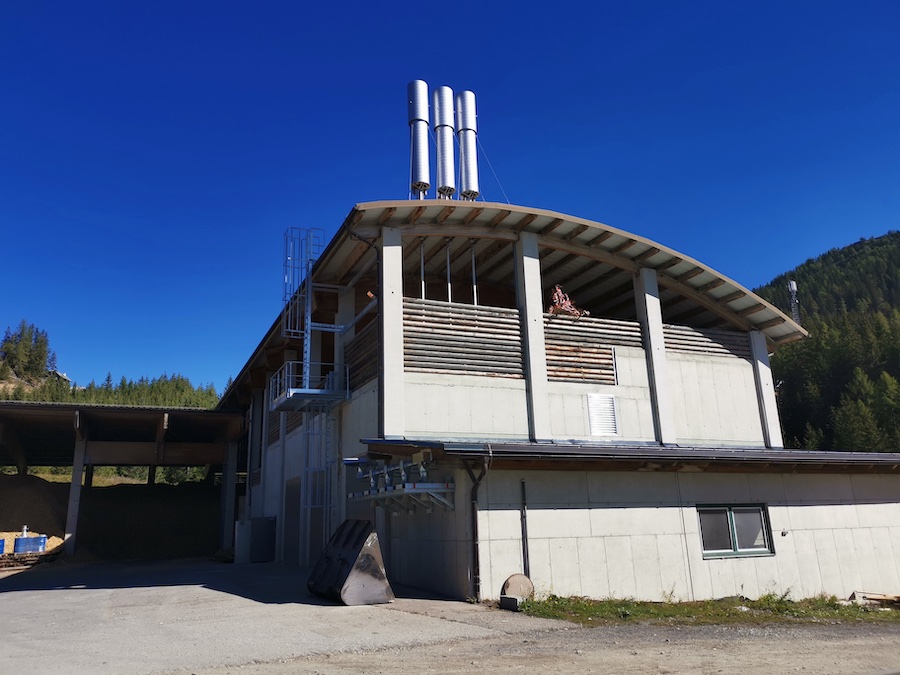 Industriegebäude mit drei metallenen Röhren auf dem Dach, offener Balkon mit Holzverkleidung und Wald im Hintergrund.