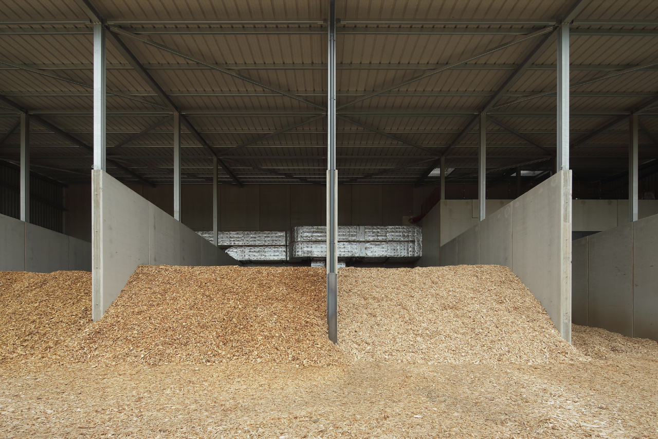 Große Holzspäne-Haufen in einer Lagerhalle, durch Betontrennwände geteilt, Stahlstützen und Dachkonstruktion sichtbar.