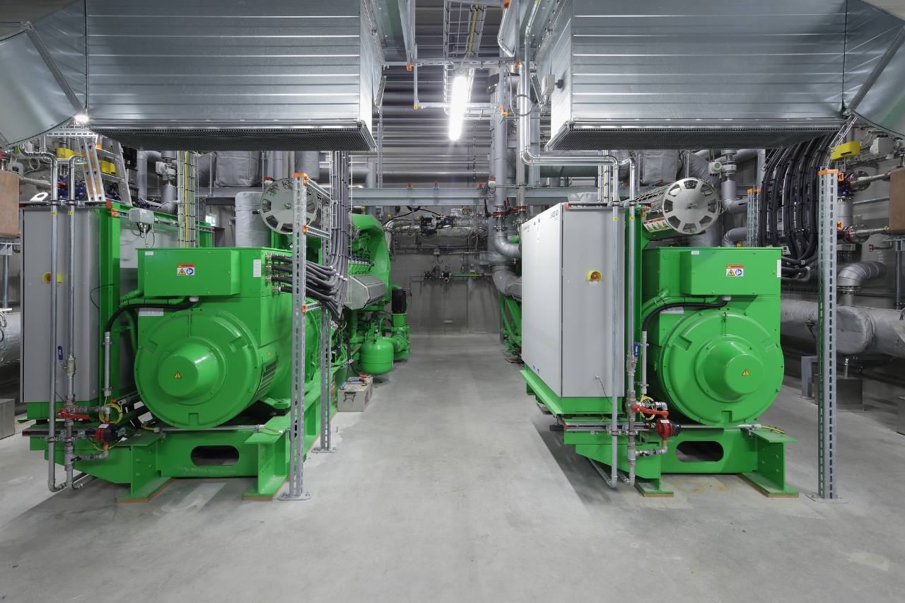 Industrieller Maschinenraum mit zwei großen grünen Generatoren, Rohrleitungen, Metallträgern, Lüftungskanälen und weißem Schaltschrank.